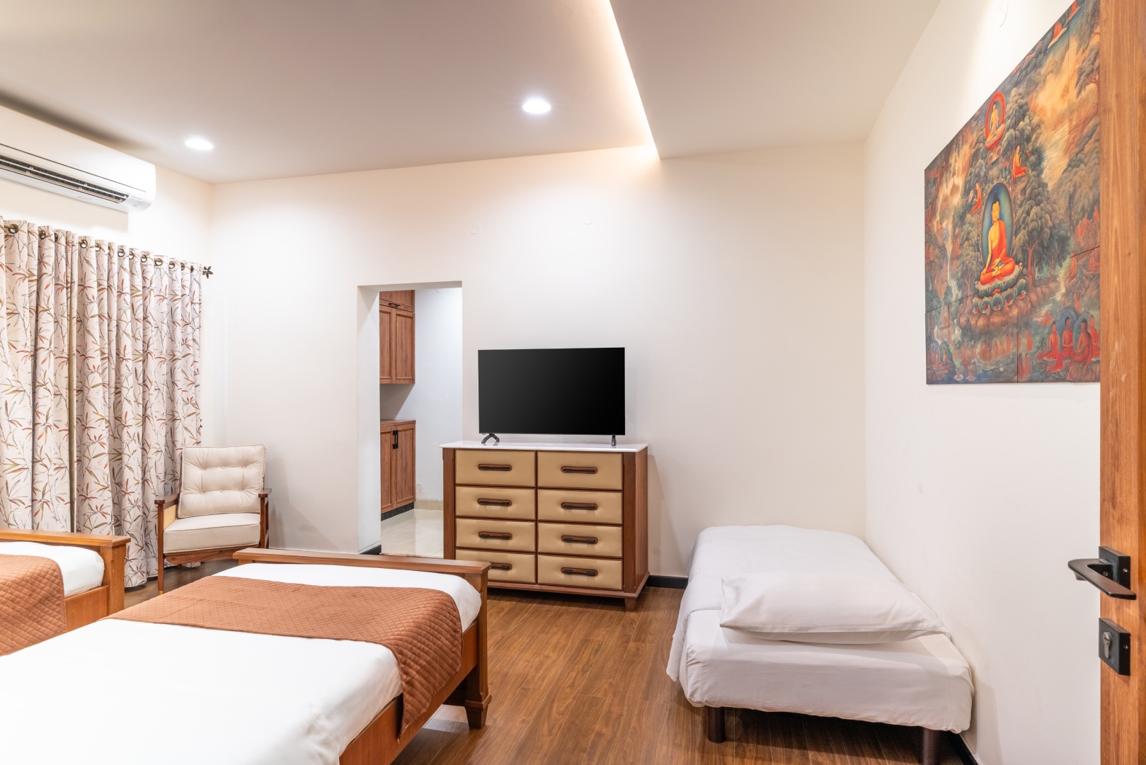 A spacious guest bedroom at Sri Narayana Vanam farmhouse featuring a combination of twin beds and an extra floor mattress, a large flat-screen TV on a contemporary dresser, and vibrant Buddhist-inspired wall art. Located at Survey number 129, Moinabad Mand