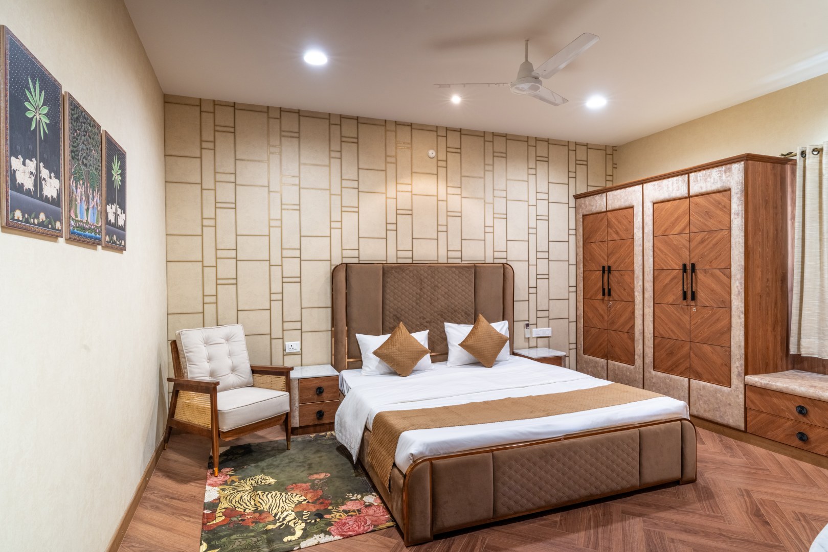 A luxury guest bedroom at Sri Narayana Vanam farmhouse featuring a king-size bed with a tufted chocolate-brown headboard, geometric wall paneling, and a large custom wooden wardrobe. The room includes a classic cream armchair and botanical wall art, located at Survey number 129, Moinabad Mandal, Aziznagar, Hyderabad, Telangana 500075. This elegantly furnished accommodation is perfect for high-end staycations and vacation rentals.