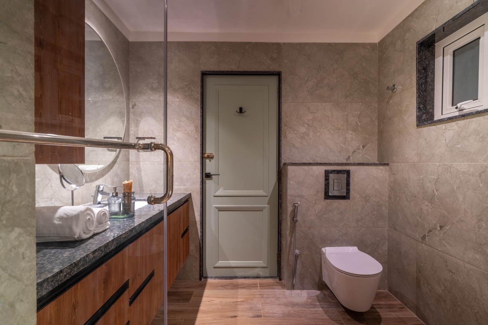 A sleek and modern ensuite bathroom at Sri Narayana Vanam luxury farmhouse, featuring stone-textured grey wall tiles, a floating wooden vanity with a dark granite top, and a large circular backlit mirror. The room includes a wall-mounted toilet and a glass
