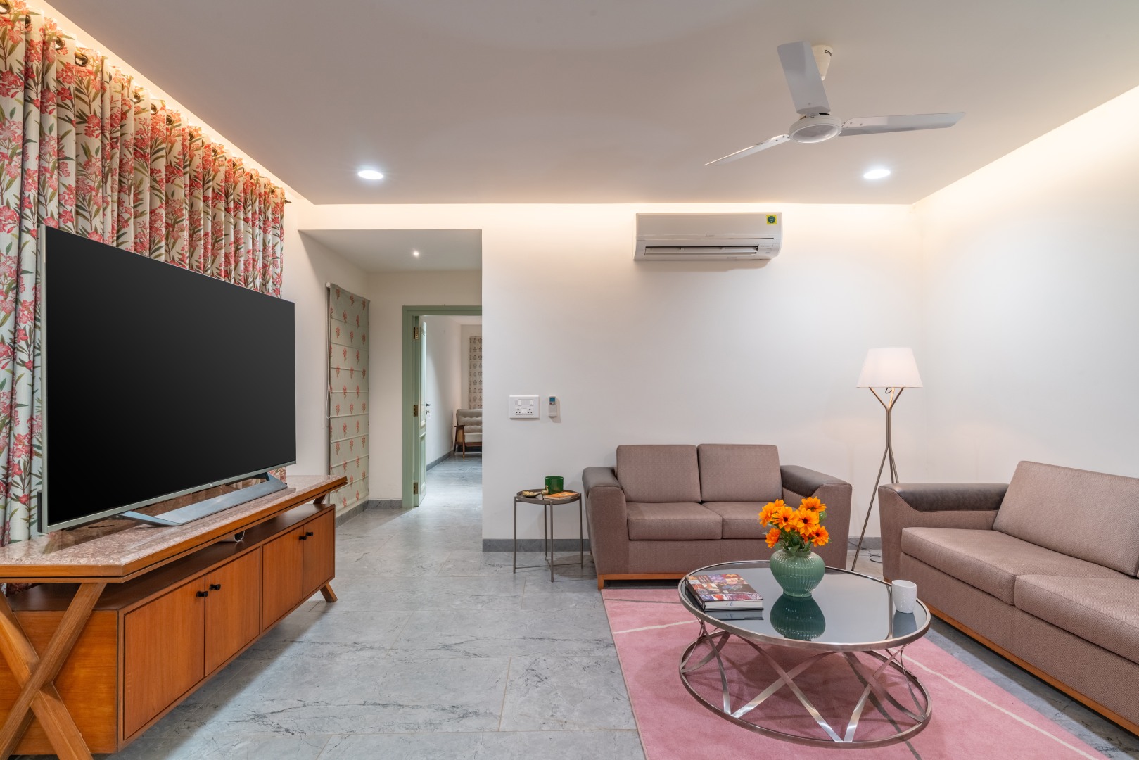 Modern entertainment lounge at Sri Narayana Vanam farmhouse in Moinabad, Hyderabad, featuring a large flat-screen TV on a wooden console, comfortable grey sofas, a glass coffee table with fresh flowers, and air conditioning at Survey number 129, Aziznagar,