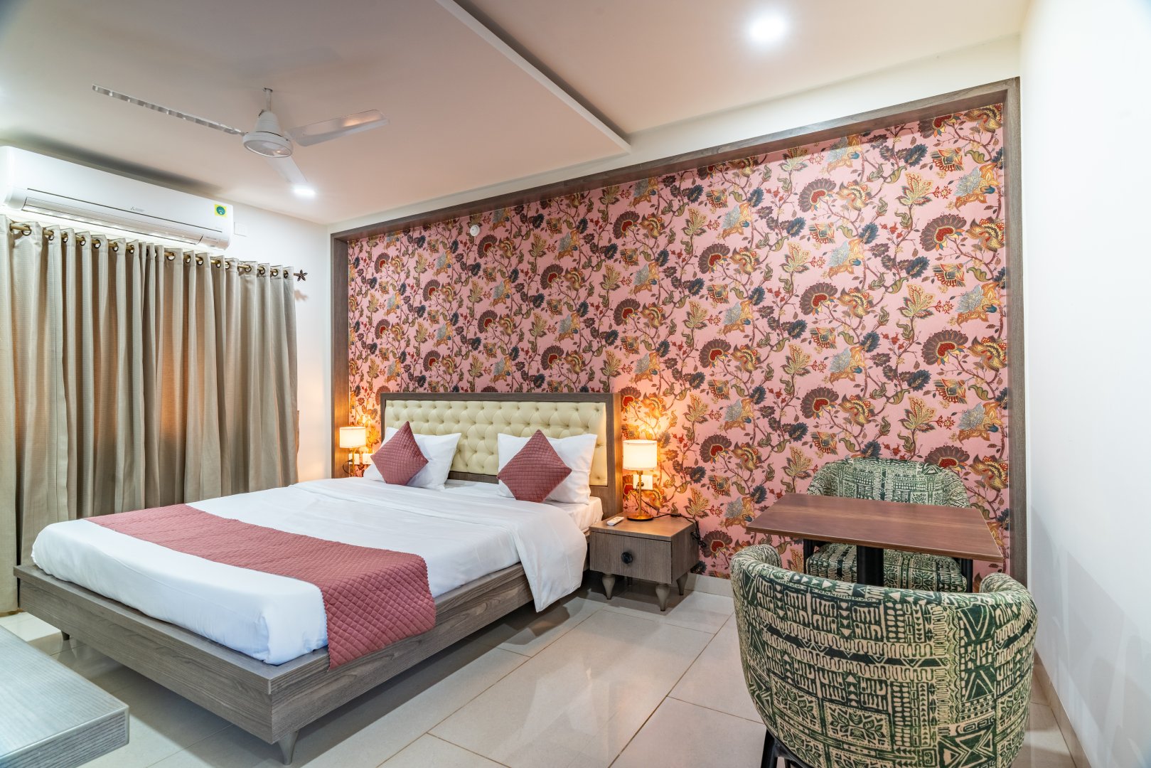 A luxury guest bedroom at Sri Narayana Vanam farmhouse featuring a king-size bed with a tufted cream headboard, vibrant pink floral-patterned wallpaper, and a cozy seating area with two patterned armchairs and a small wooden table. Located at Survey number