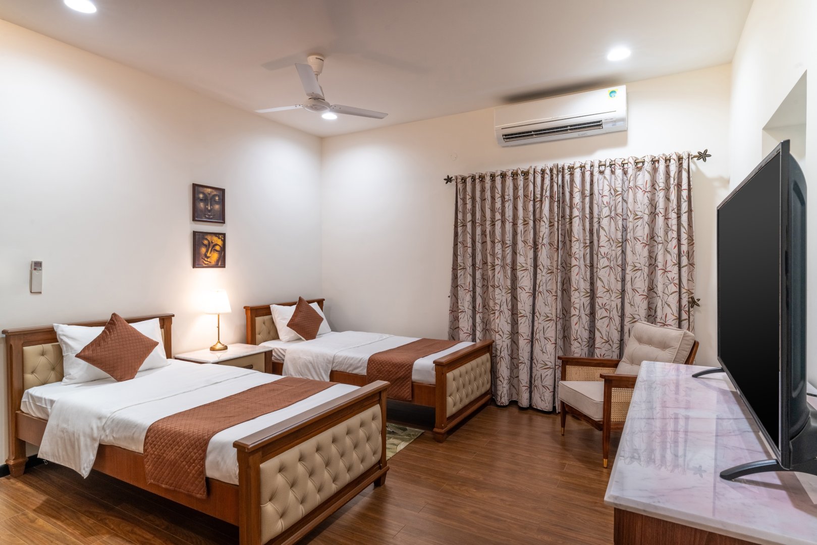 A comfortable guest bedroom at Sri Narayana Vanam farmhouse featuring twin beds with tufted cream headboards and brown accents. The room includes a flat-screen TV on a marble-top console, a classic armchair, and bamboo-patterned curtains. Located at Survey