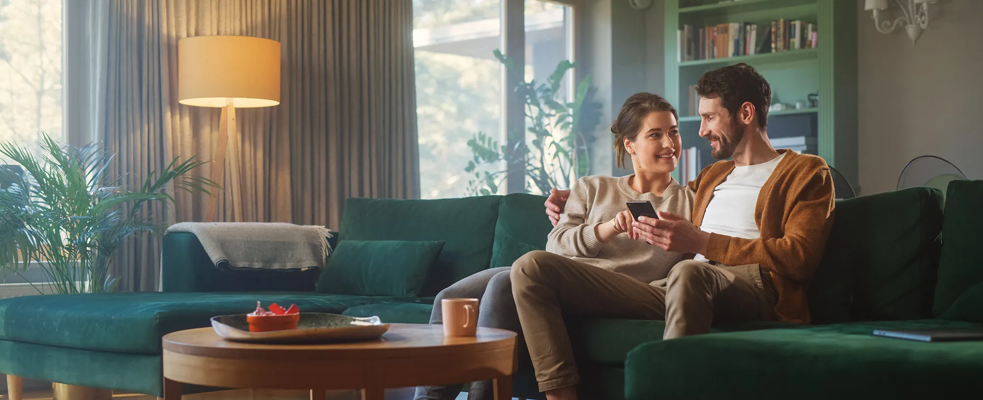 Paar sitzt auf einem grünen Sofa in einem gemütlichen Wohnzimmer und schaut gemeinsam auf ein Smartphone.