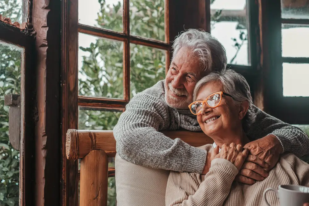 Älteres Paar sitzt an einem Fenster, der Mann umarmt die Frau liebevoll von hinten.