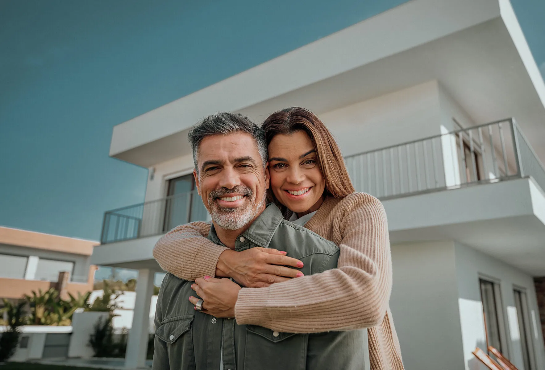 Ein glückliches Paar umarmt sich vor einem modernen, zweistöckigen Haus bei blauem Himmel.