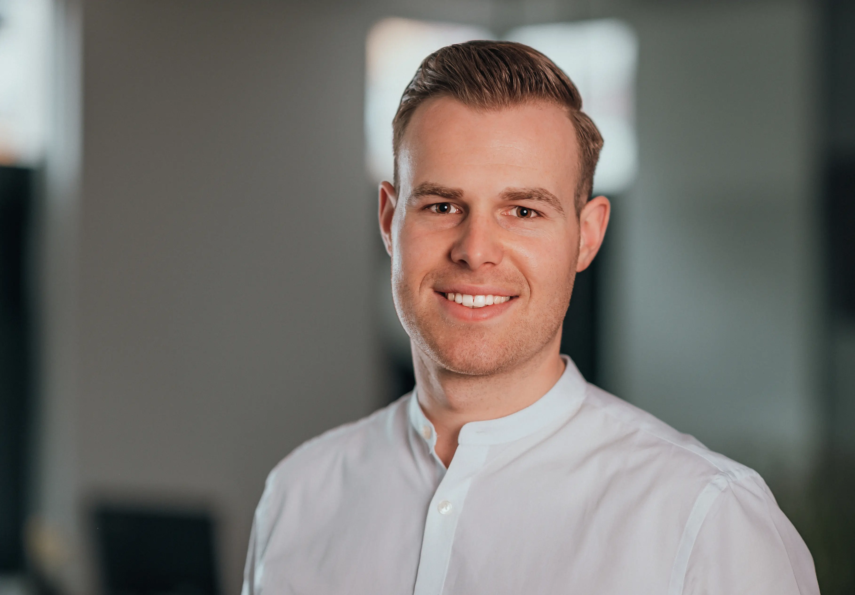 Junger Mann mit hellbraunen Haaren, der in einem weißen Hemd lächelnd in die Kamera schaut.