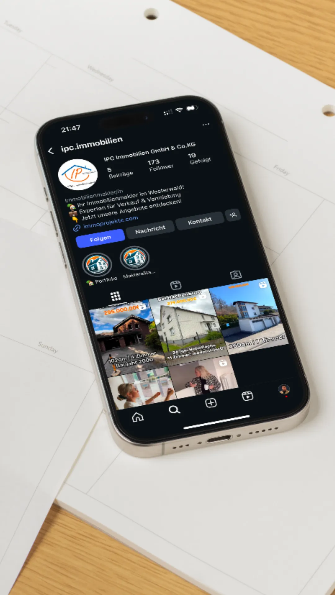 Smartphone zeigt Instagram-Profil von ipc.immobilien mit Immobilienangeboten auf einem Schreibtischkalender.