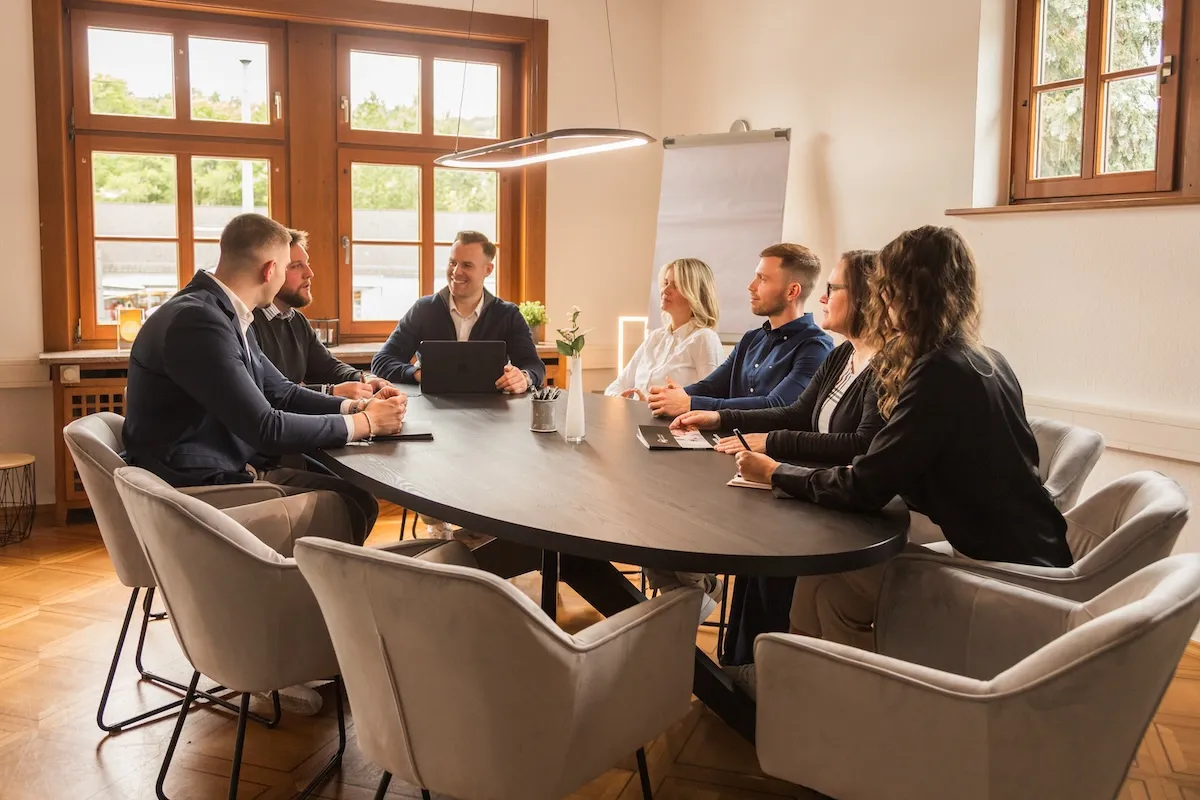 Sieben Personen sitzen um einen ovalen Konferenztisch in einem hellen Raum mit Holzfenstern und führen eine Besprechung.