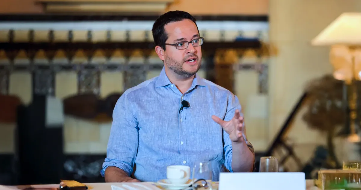 A man in a blue shirt speaks animatedly, sitting at a table with a coffee cup and papers. The background features elegant decor, fostering a professional tone.