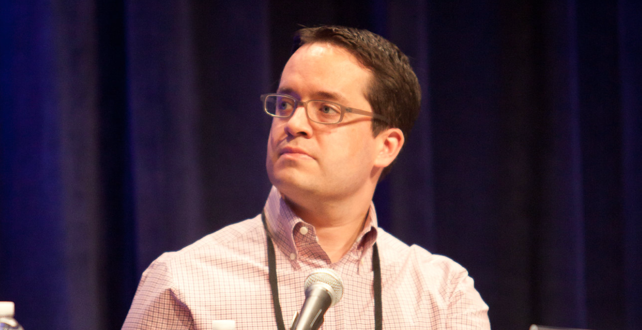 Rugby players in orange and black jerseys engage in an intense scrum on a grassy field, with dust rising around them, conveying energy and determination.