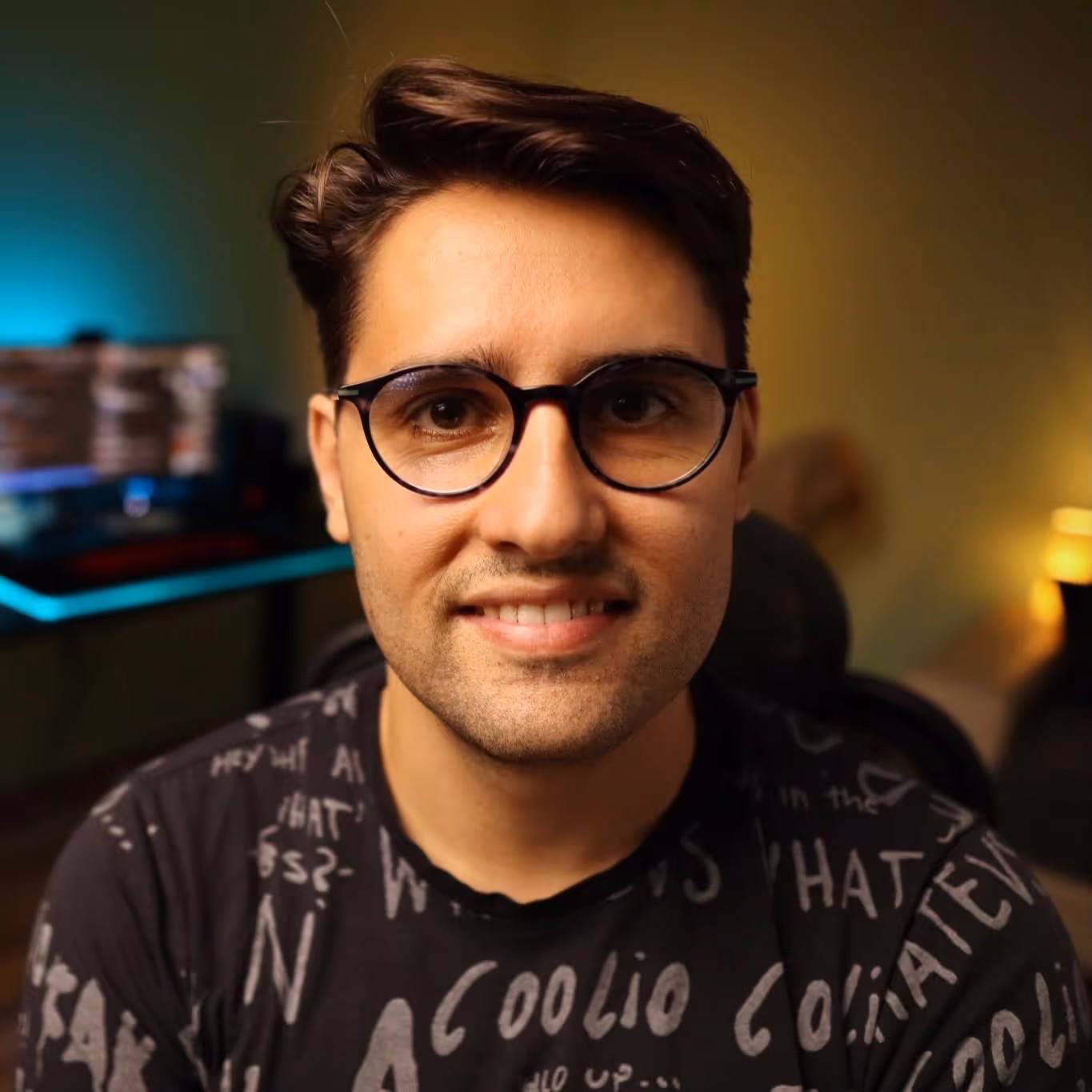 Man wearing glasses with dark hair, smiling slightly, in an indoor setting with blurred background.