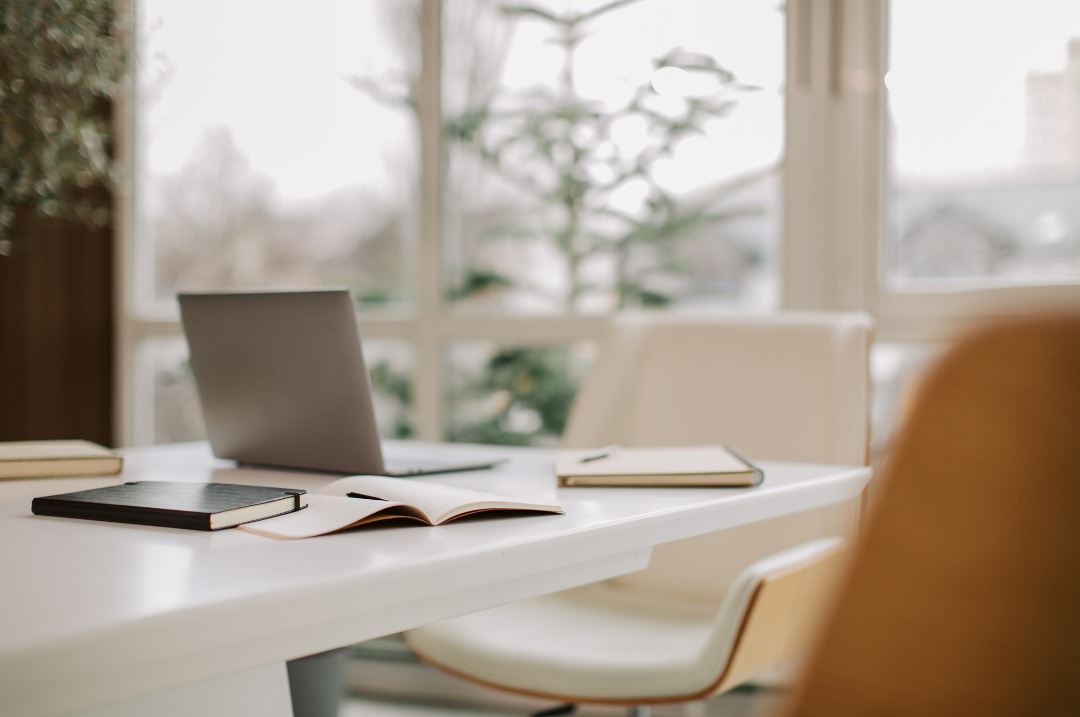 Image d'un bureau de travail en pleine journée près d'une fenêtre, sur lequel se trouve des carnets de notes et un ordinateur
