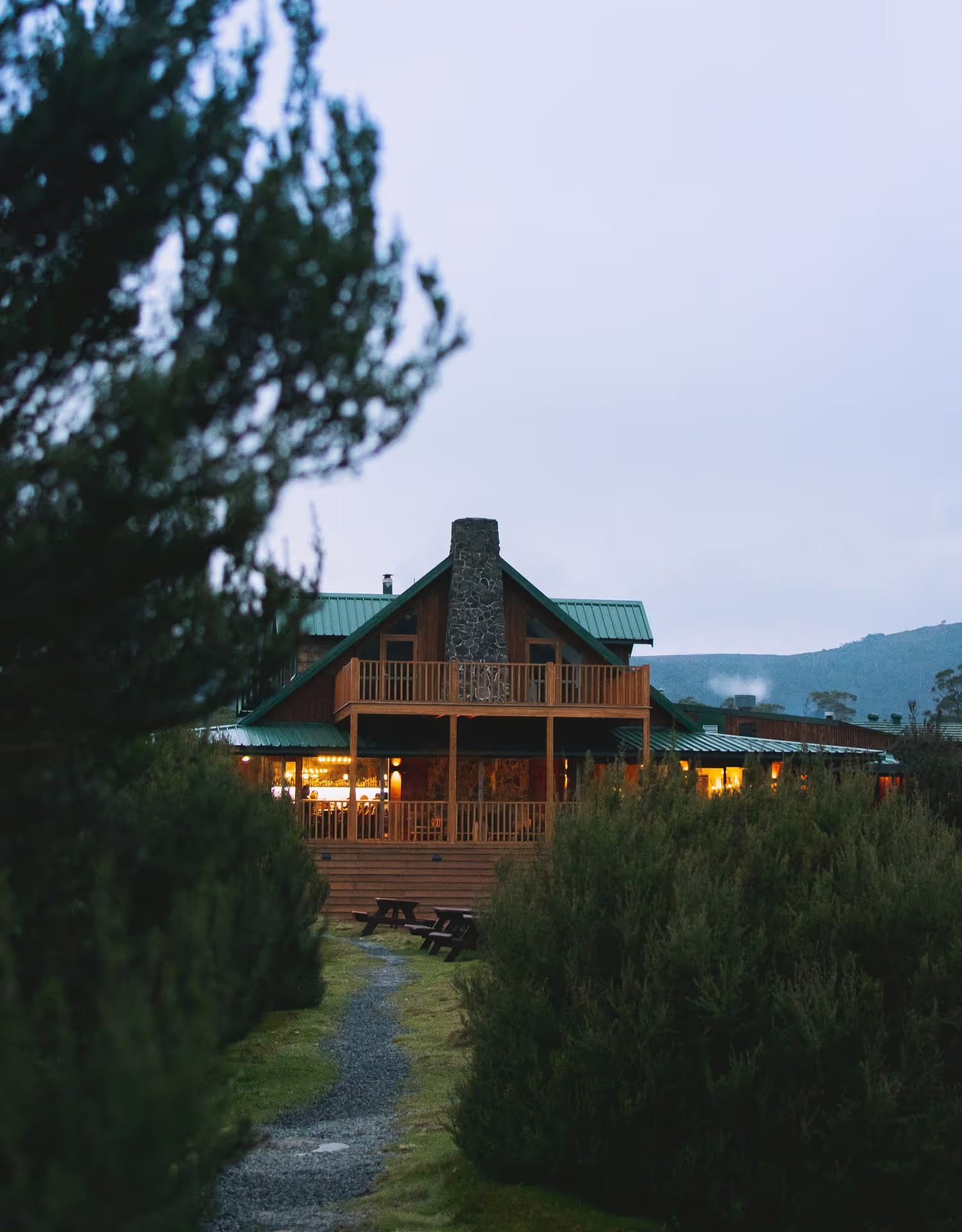 A rustic wooden cabin with a stone chimney, warmly lit in the evening, surrounded by greenery and a winding gravel path.