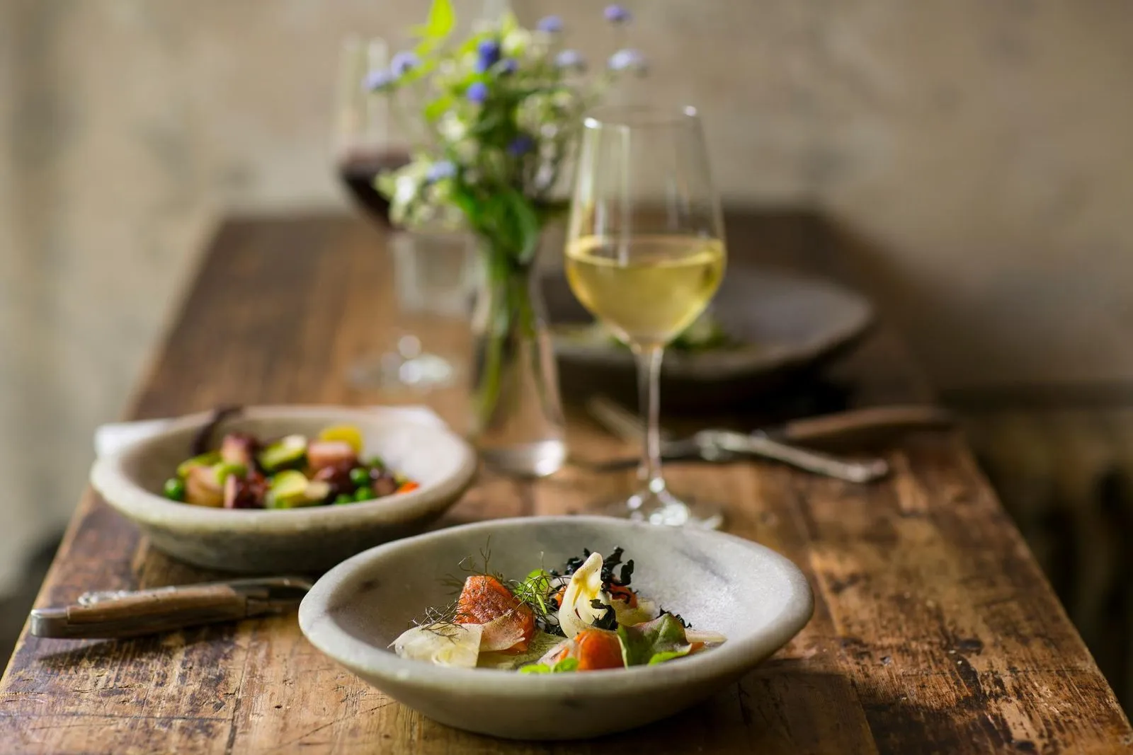A beautifully plated dish with seafood and greens beside a fresh salad, wine glasses, and a small vase of flowers on a rustic wooden table.