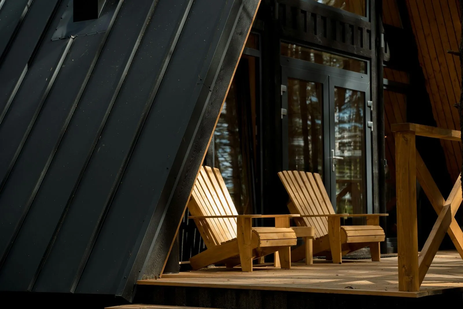 Sunlit wooden Adirondack chairs on a deck, framed by an angled black metal roof and large glass doors, amidst a serene forest backdrop.