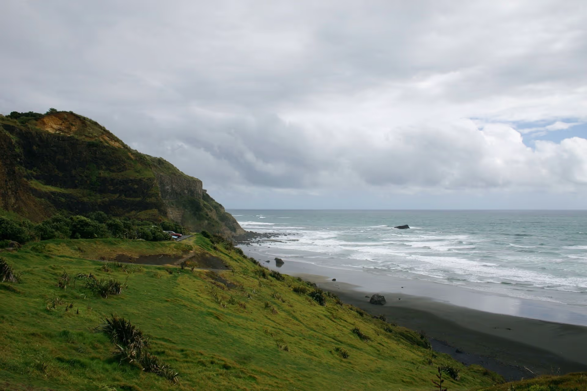 A scenic coastal view with grassy cliffs meeting the ocean under a cloudy sky, showcasing waves lapping at the dark sandy beach below.