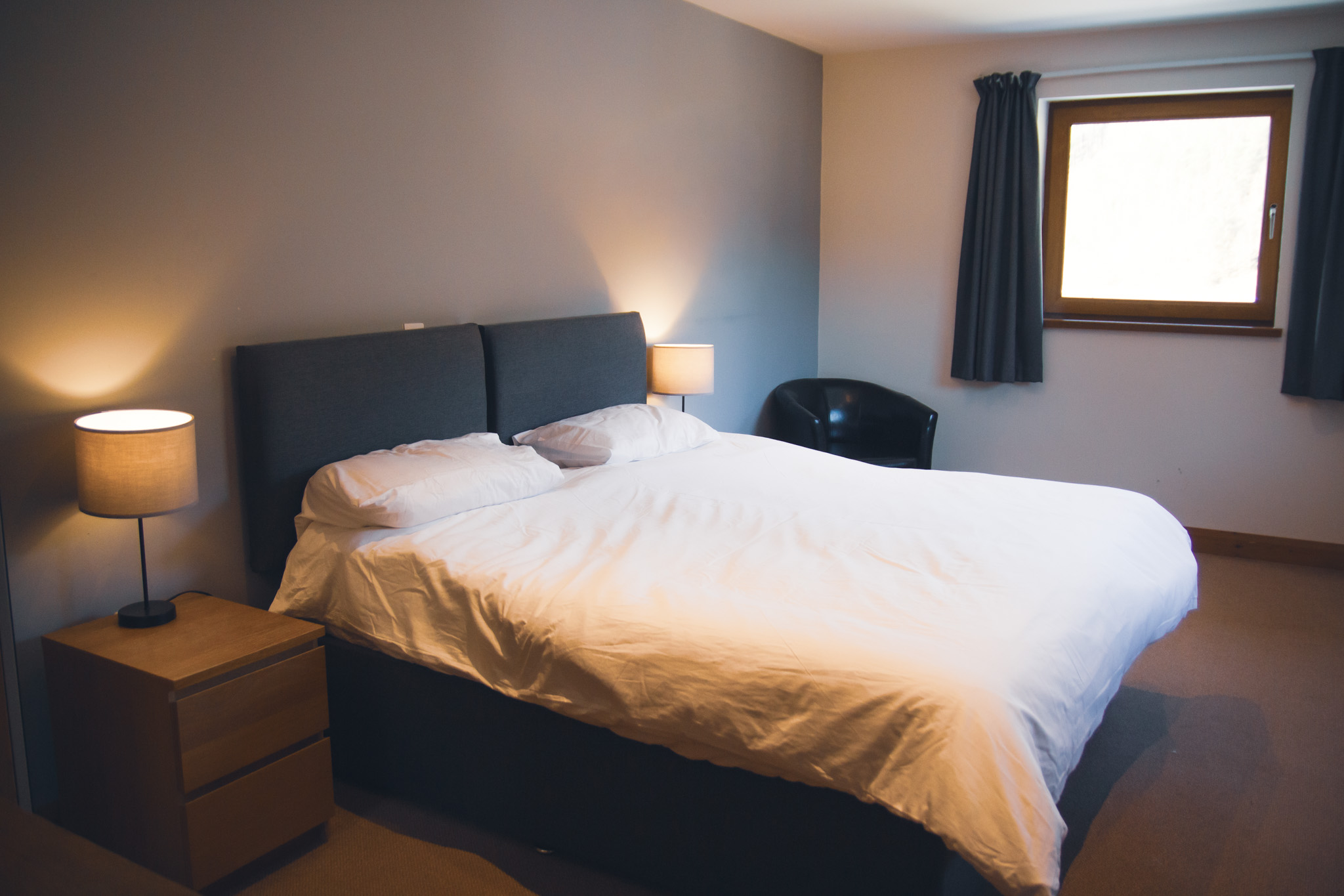 Cozy, modern living room with angled wood beams, large windows, a light gray sofa, and a decorative glass coffee table. Greenery visible outdoors.