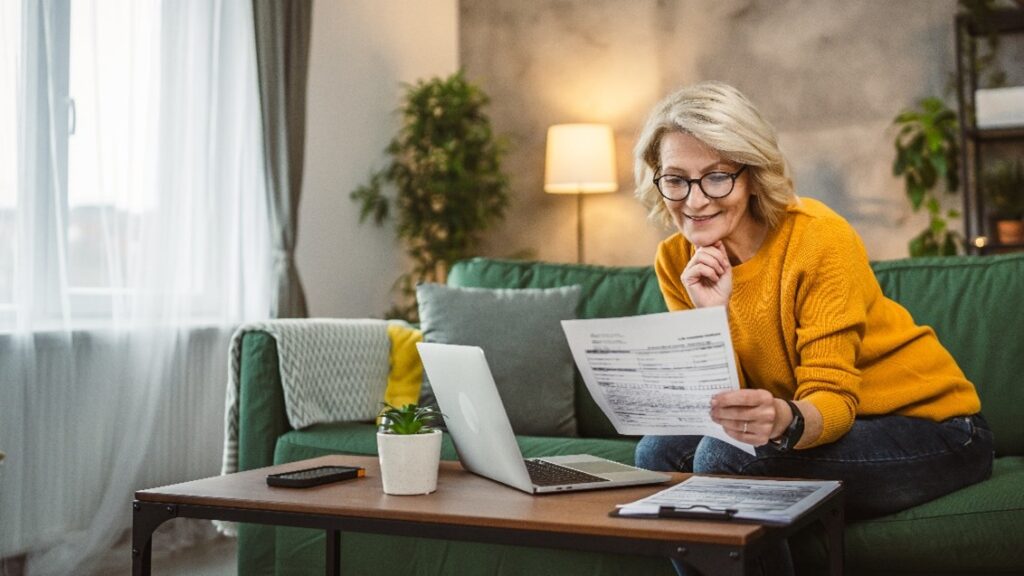 Person in a yellow sweater sitting on a green couch, reviewing documents with a laptop on the coffee table in the U.S.