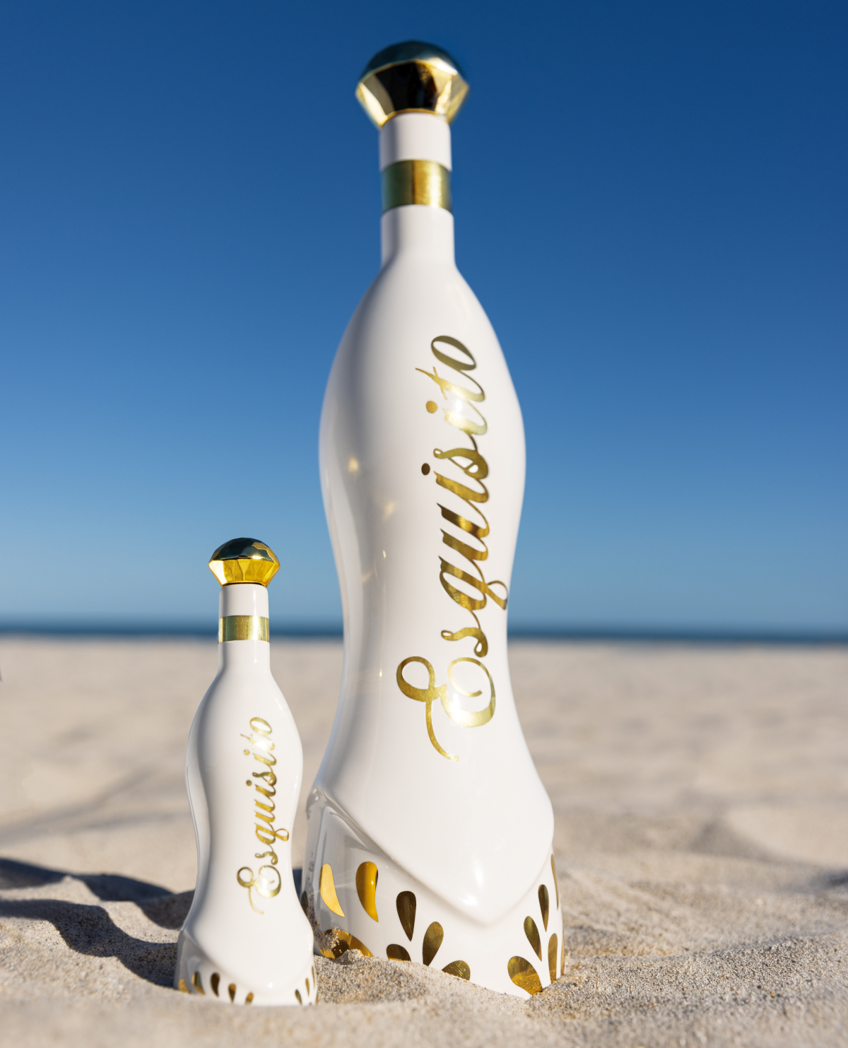 Two white Esquisito Tequila bottles with gold accents standing upright on sand against a clear blue sky.