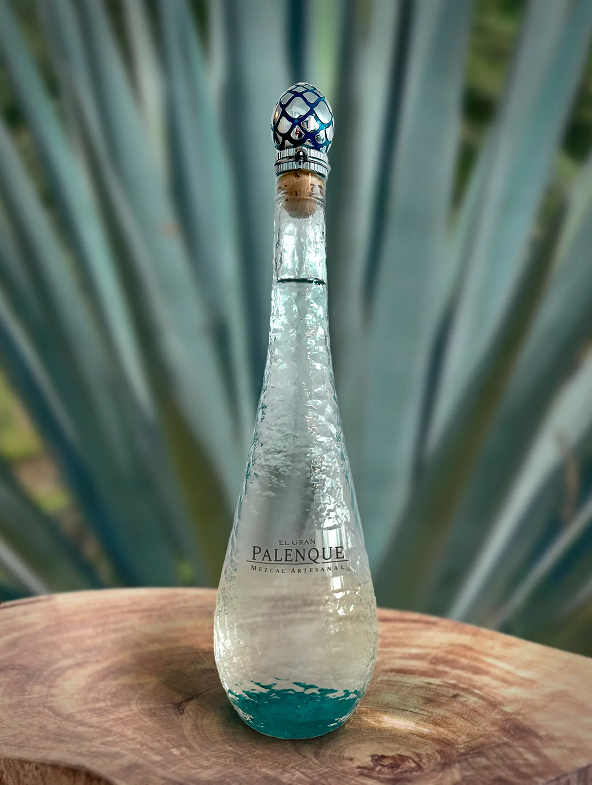 Tall textured glass bottle of El Gran Palenque Mezcal Artesanal with decorative stopper, placed on a wooden surface with blurred agave plant in background.