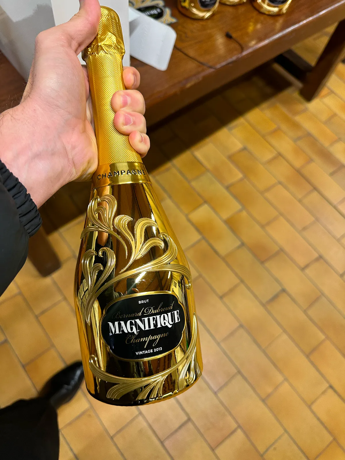 Hand holding a shiny gold bottle of Bernard Dubreuil Magnifique Brut Champagne vintage 2013 with decorative embossed design.
