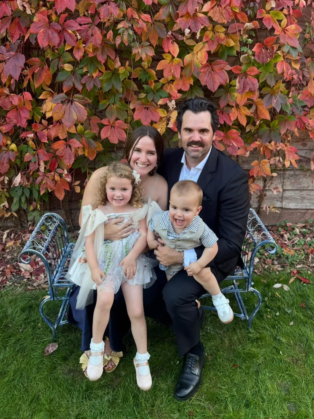 Madeline and her family of four sitting on a metal bench outdoors with autumn red and orange leaves in the background.