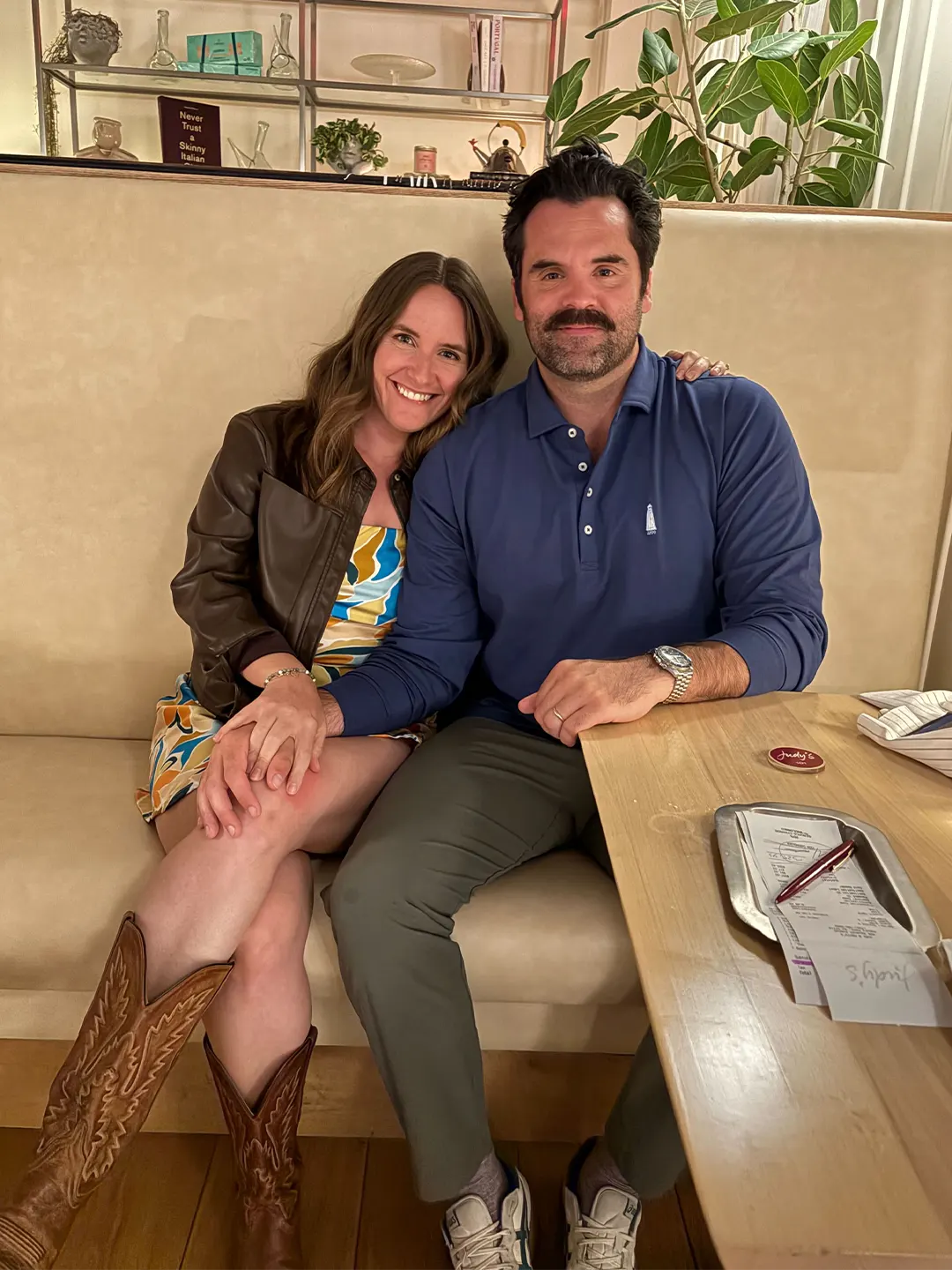 Madeline and her husband sitting on a beige booth in a restaurant, holding hands, with plants and shelves in the background.