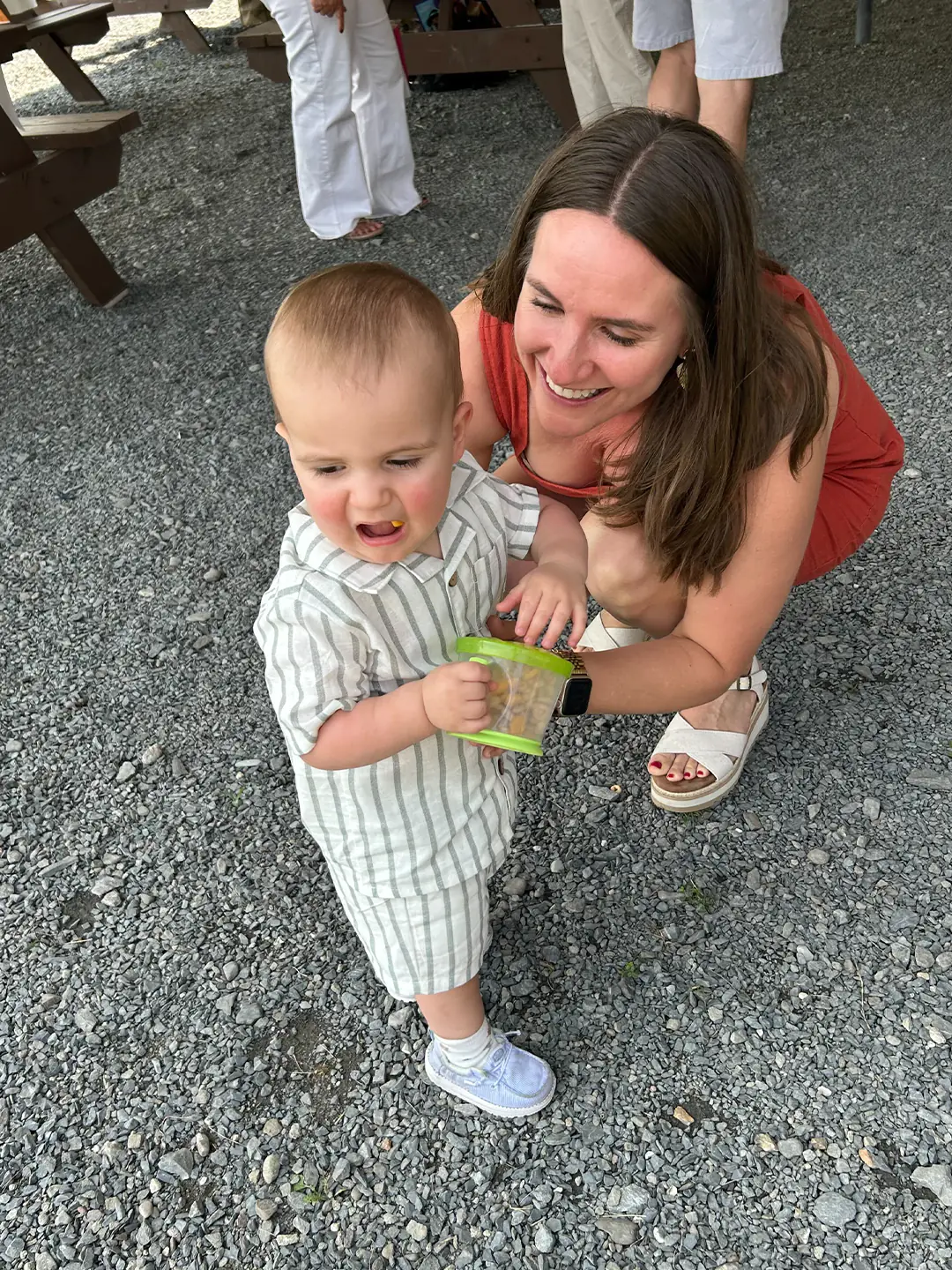 Madeline crouches beside her toddler holding a green container, standing on gravel outdoors.