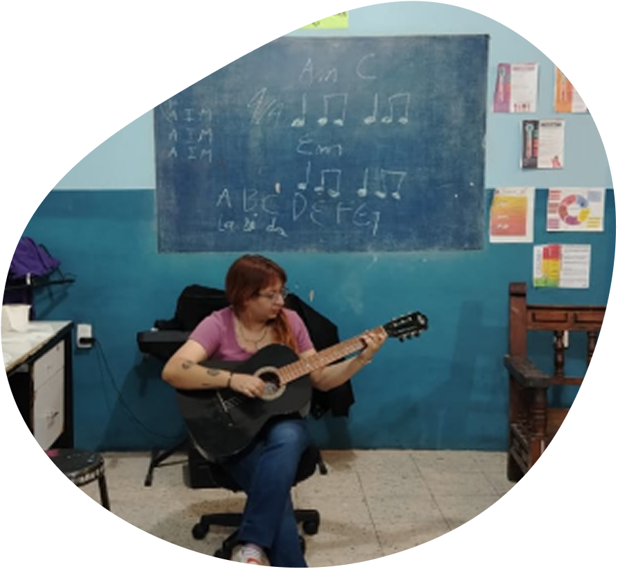 Person sitting on a chair playing a black acoustic guitar in a room with blue walls and a chalkboard showing musical notes and letters.