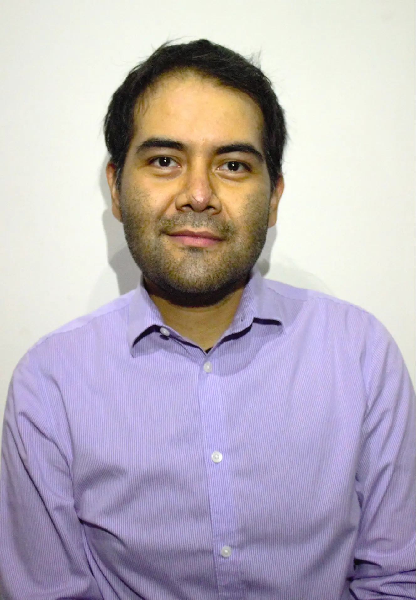 Man with short dark hair wearing a light purple button-up shirt, smiling slightly against a plain white background.