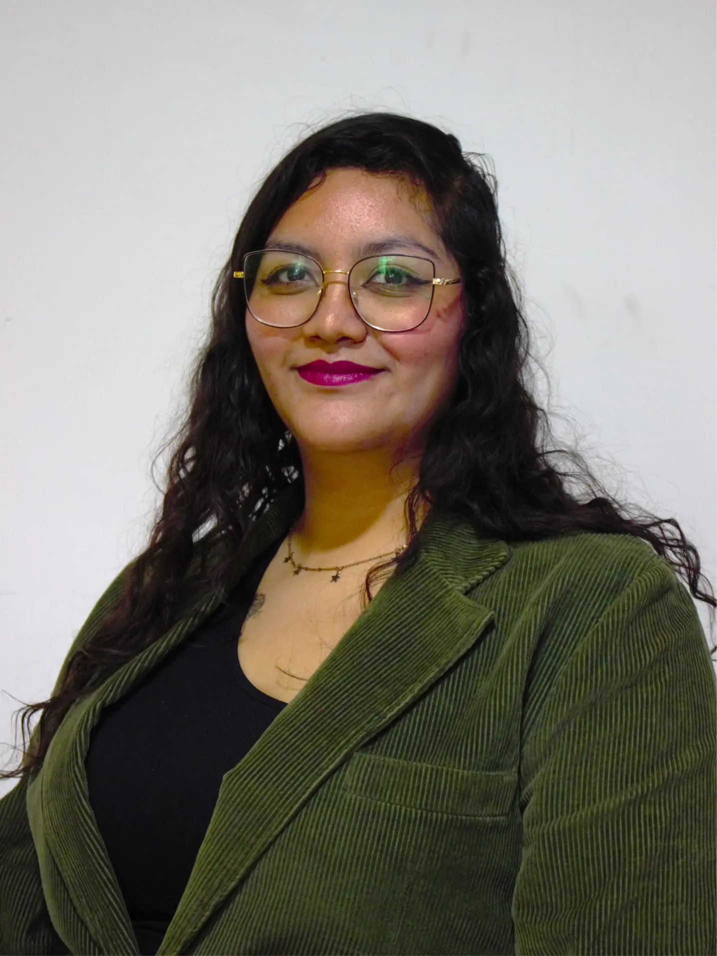 Woman with long curly hair wearing glasses, a green corduroy blazer, black top, and a star necklace against a plain light background.