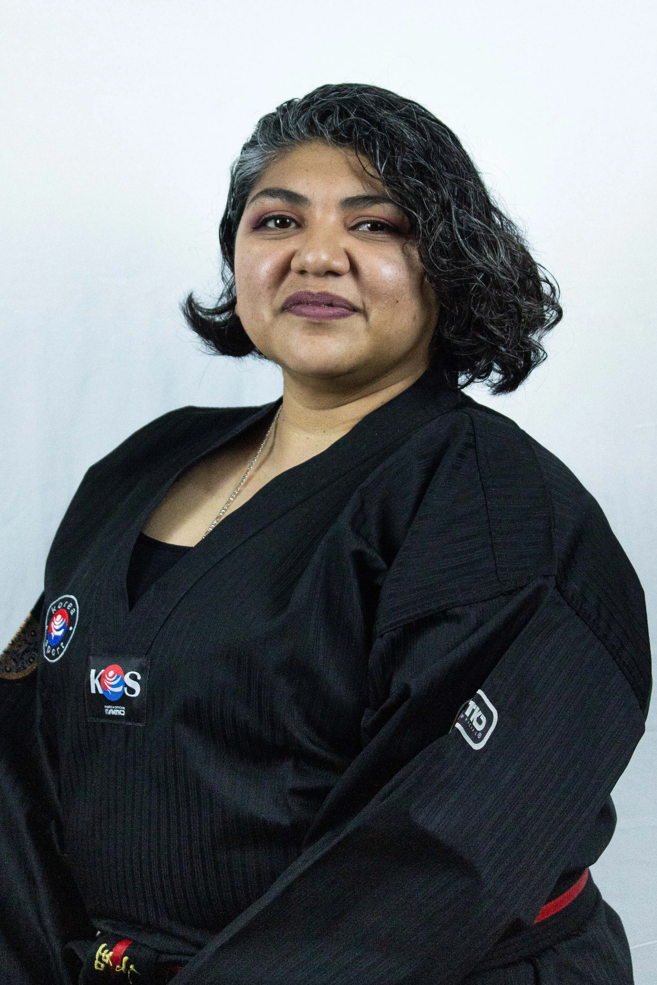 Person with short curly hair wearing a black martial arts uniform with patches on the chest and sleeve, against a plain light background.