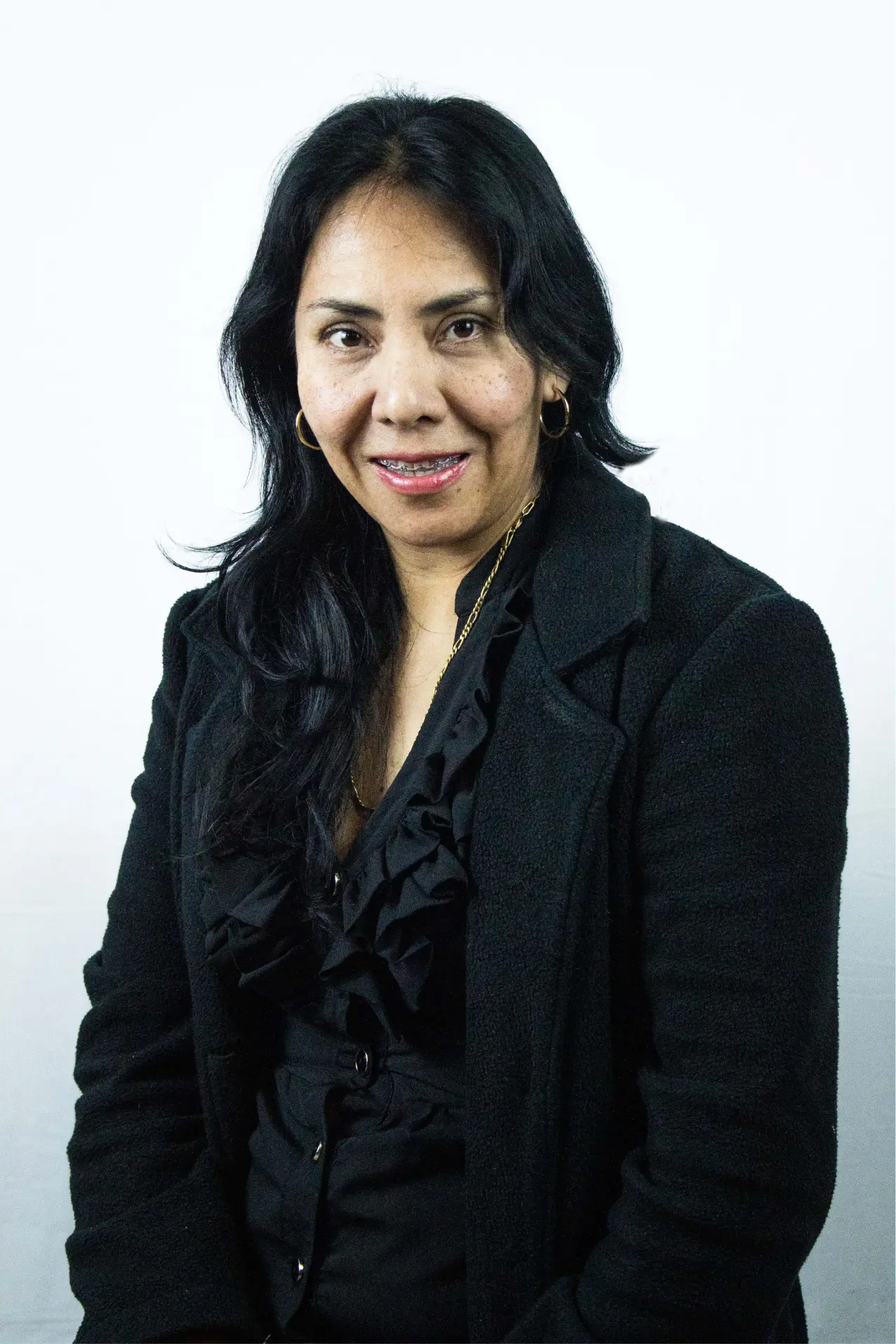Woman with long black hair wearing a black coat and ruffled blouse, smiling slightly against a plain white background.