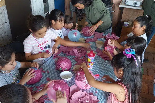 Children and an adult crafting papier-mâché objects at a table covered with a blue checkered cloth.