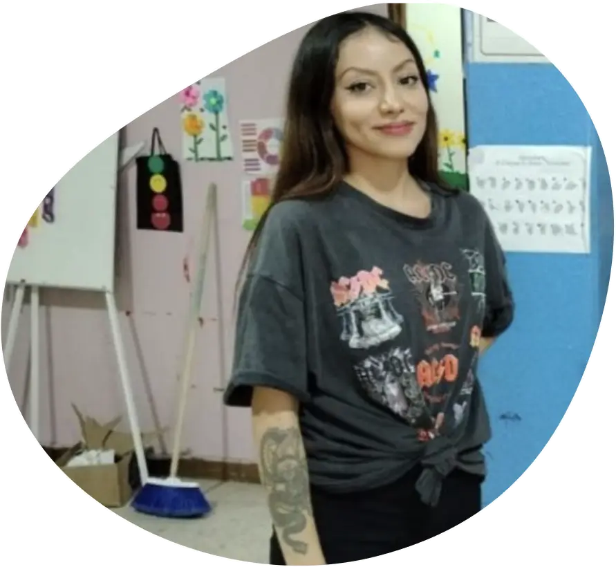 Young woman with long dark hair and tattoo on her arm, wearing a dark graphic t-shirt tied at the waist, smiling indoors with colorful wall drawings and a broom in the background.