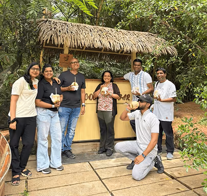 Six people enjoying drinks outdoors in front of a small hut with a thatched roof in a green garden setting.
