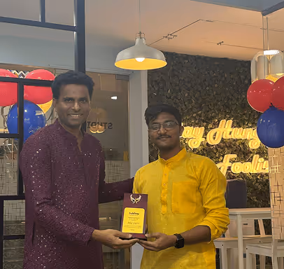 Two men in traditional attire holding a plaque inside a decorated room with balloons and a sign in the background.