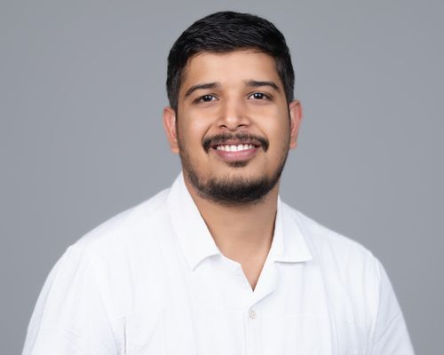 Smiling man with short black hair and beard wearing a white collared shirt against a gray background.