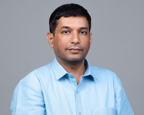 Man with short black hair wearing a light blue shirt posing against a plain gray background.