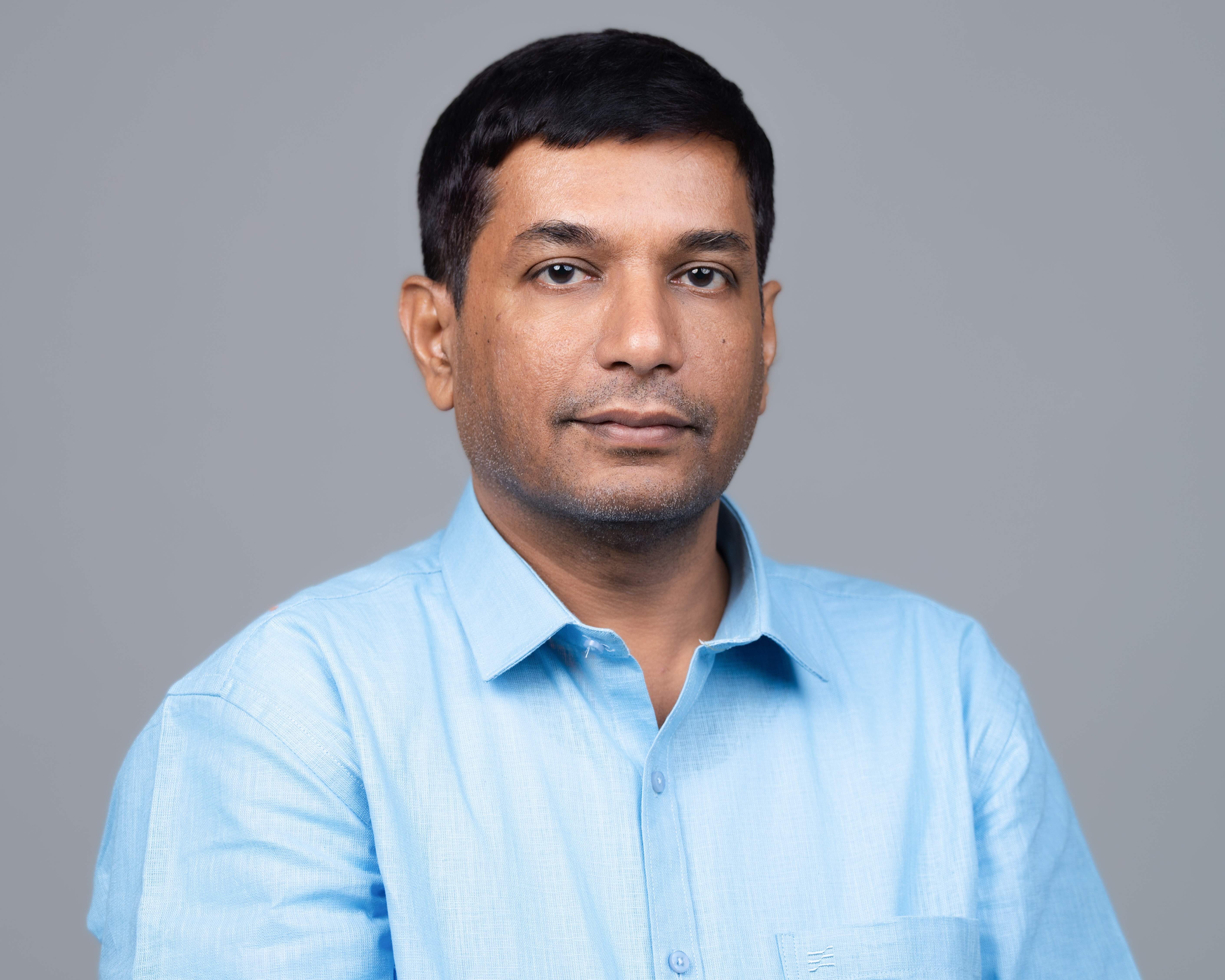 Man with short black hair wearing a light blue shirt posing against a plain gray background.