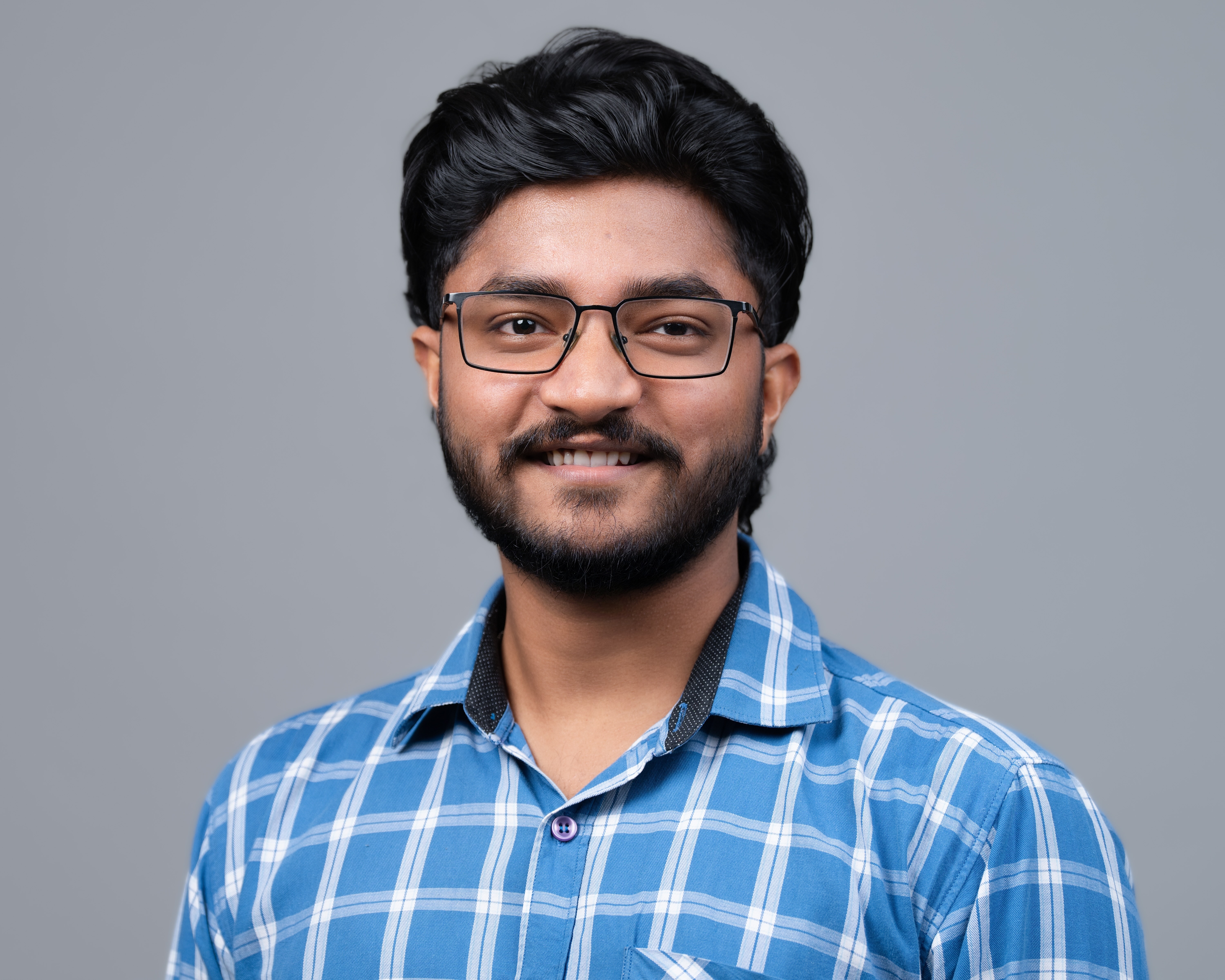 Young man wearing black-rimmed glasses and a blue checkered shirt smiling against a gray background.