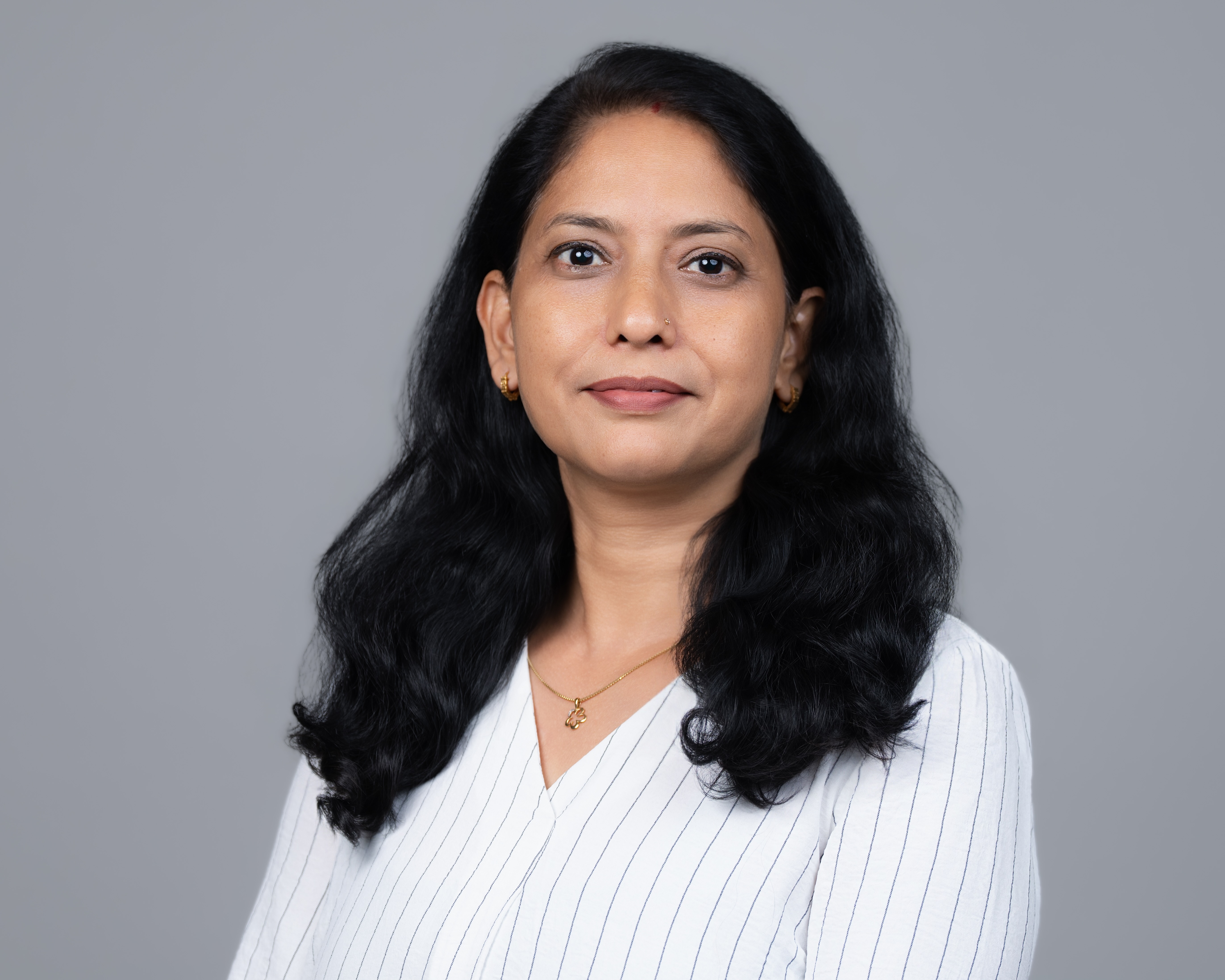 Portrait of a woman with long black hair wearing a white pinstriped blouse and gold jewelry, against a plain gray background.