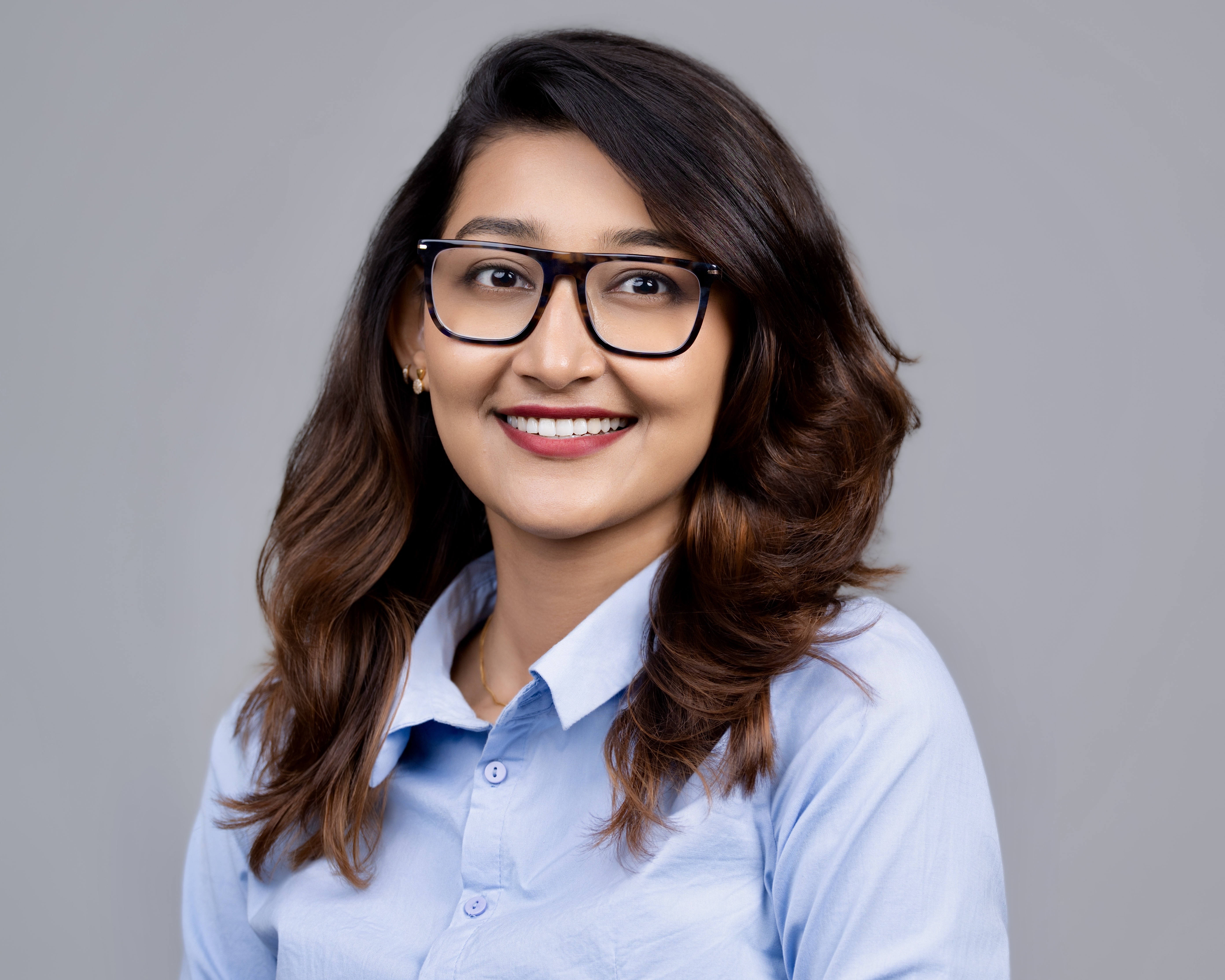 Smiling woman with long brown hair, wearing glasses and a light blue button-up shirt against a gray background.