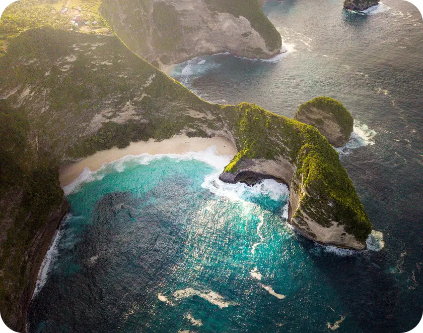 Aerial view of a curved cliff covered with greenery surrounding a turquoise bay with white sandy beach and waves crashing.