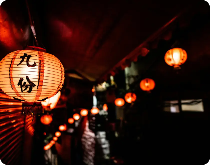 Glowing orange paper lanterns hanging in a dark alley, with one lantern featuring black Chinese characters.