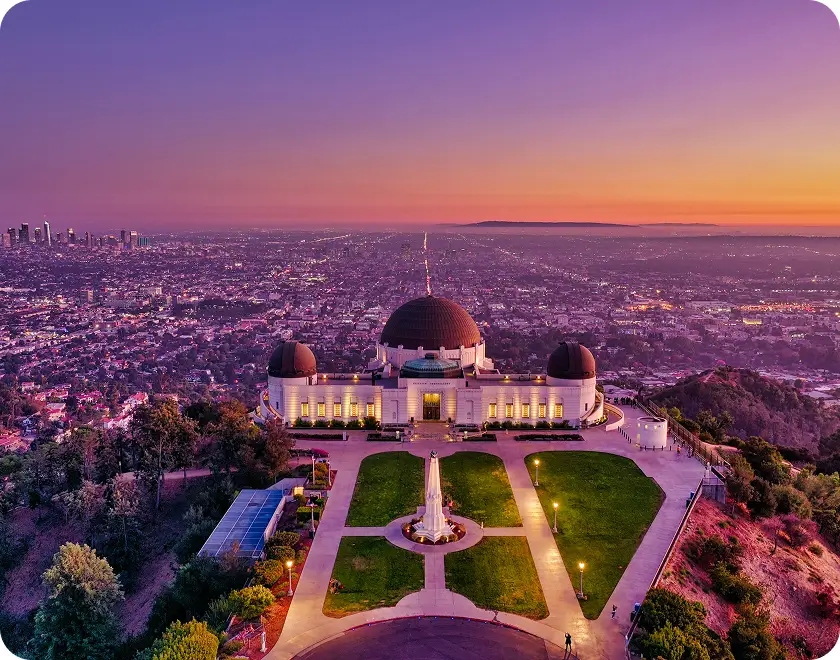 Griffith Observatory overlooking Los Angeles at sunset with city lights and purple-orange sky.