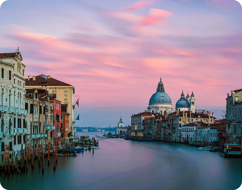 Venice canal at sunset with historic buildings lining both sides and a large domed church in the background under a pink and purple sky.