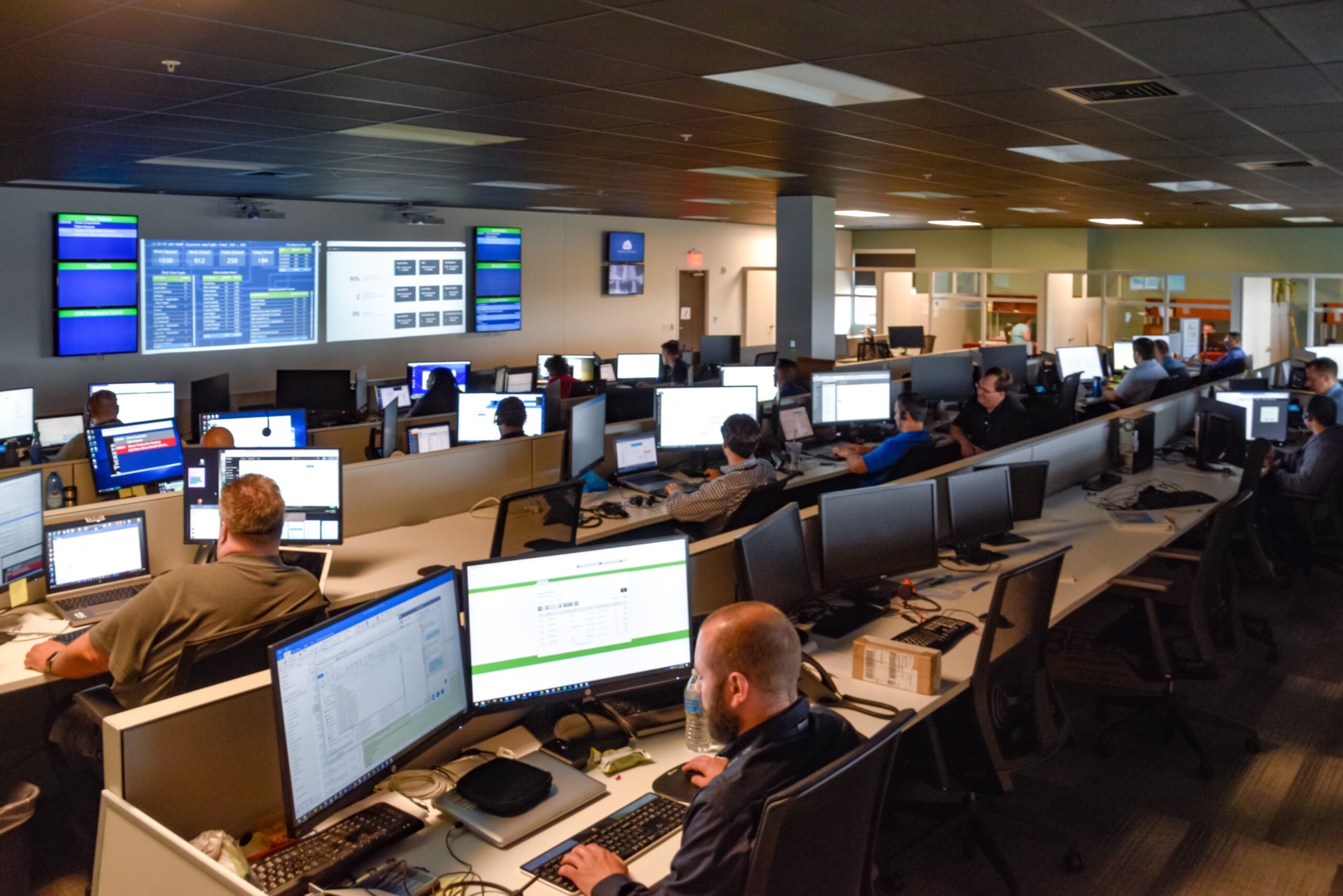 Fortress SRM security operations centre floor with analysts at multi-monitor workstations and a wall of monitoring dashboards