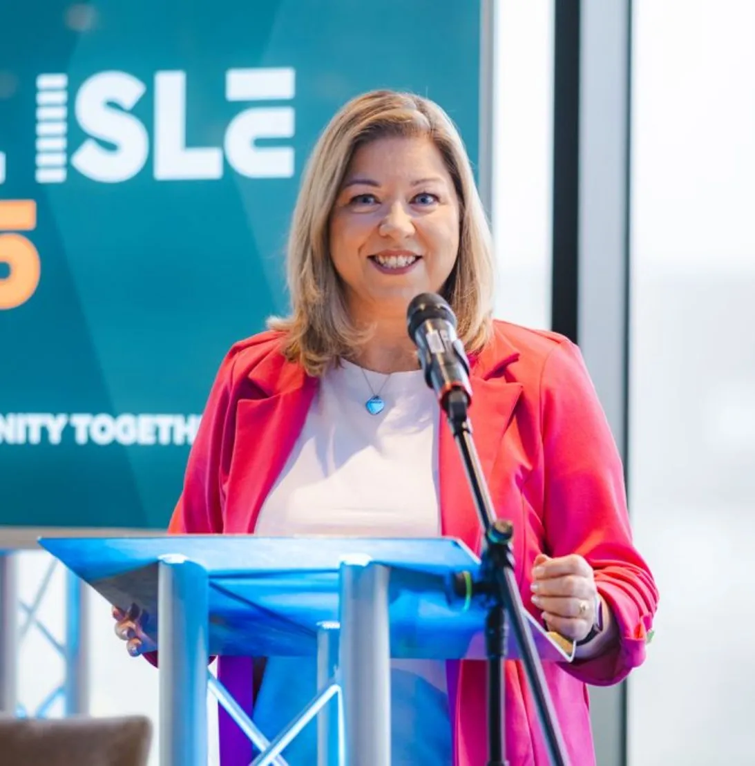 Woman in a pink blazer speaking at a podium with a microphone in front of a screen.