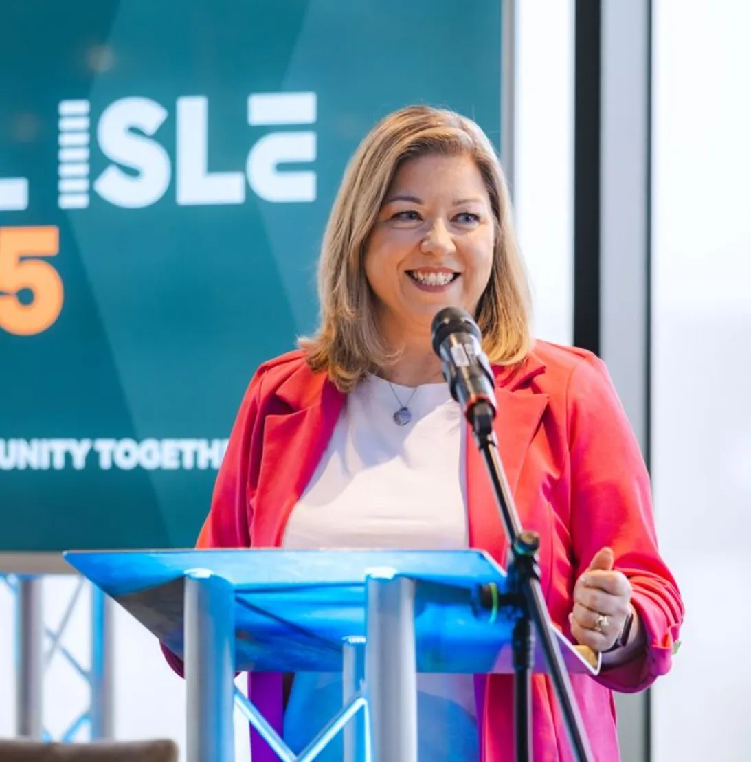 Smiling woman in a pink blazer speaking at a podium with a microphone against a background screen.